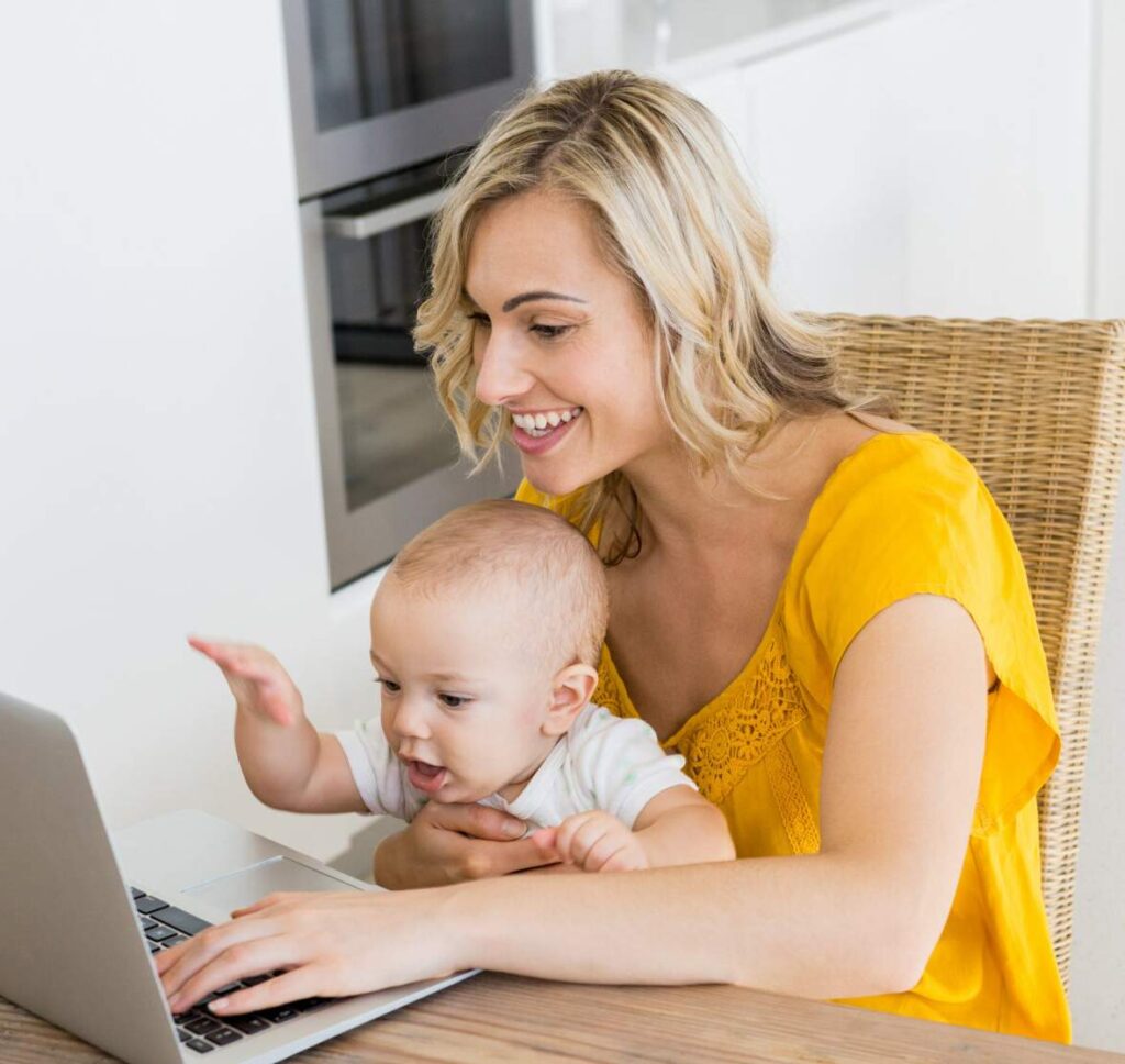 A young mother writes on her computer while balancing a baby on her lap. 